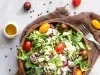 Fotografia de uma salada em uma tigela de madeira, repleta de folhas verdes, tomates-cereja coloridos e queijo branco esfarelado, com um toque de molho balsâmico. Um pequeno recipiente de azeite e tomates inteiros decoram a cena.