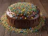 Fotografia de um bolo de chocolate com um brilho elegante, com glacê escorrendo pelas laterais. Decorado com uma variedade de confeitos coloridos, a imagem destaca o bolo em um fundo de madeira.