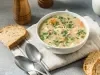 Imagem de um bowl branco com a receita de canja de galinha decorada com salsinha. Ao lado, pedaços de pão e duas colheres.