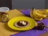 Fotografia de uma mesa de madeira com dois panos, roxo e amarelo, um por cima do outro. Apoiado neles tem um prato amarelo com uma panqueca de banana e creme de avelã. Ao redor tem uma lata de NINHO Nutri Advance, um cacho de bananas e o creme.