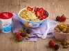 Fotografia em tons de lilás em uma bancada de madeira clara, um bowl colorido com o iogurte de abacate com frutas vermelhas e granola decorando. Ao lado, um potinho com granola e frutas espalhadas na bancada.