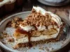 Em cima de uma mesa, fatia de banoffee com massa de biscoitos crocante, chantilly cremoso, bananas em rodelas, decorado com chantilly e nozes picadas.