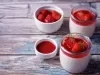 Fotografia de dois recipientes de vidro pequenos com mousse e uma calda de morangos e frutas vermelhas, sobre uma mesa de madeira. Ao redor tem dois recipientes, com morangos inteiros e o outro, com um pouco da calda.