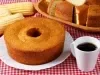 Fotografia de um bolo de milho com coco, exibido em um prato branco sobre uma toalha de mesa xadrez vermelha e branca. O bolo, com crosta dourada, é acompanhado por espigas de milho e uma caneca de café.