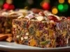 Fotografia de uma fatia de bolo de frutas, repleta de frutas secas coloridas e nozes, com um toque de açúcar em pó. O fundo apresenta luzes festivas em vermelho, verde e dourado, criando um ambiente natalino.