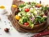 Fotografia de uma salada vibrante em uma tigela, com uma mistura de folhas verdes, tomates-cereja em várias cores e queijo branco esfarelado, regada com molho balsâmico. A composição tem um recipiente de azeite e grãos de pimenta.