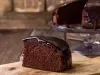 Fotografia de um bolo de chocolate fofinho com uma fatia em destaque, com uma cobertura escura e lisa. Ao fundo, um bolo está em um suporte de vidro, em um ambiente rústico.