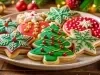 Em cima de uma mesa, prato com biscoitos em formato de árvores de Natal, estrelas, decorados com glacê colorido e confeitos coloridos de bolinhas vermelho.