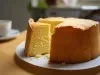 Um bolo grande e fofo de cor amarela clara, com uma fatia já cortada. Ele está sobre um prato branco, em uma mesa de madeira clara. Ao fundo, há uma xícara branca desfocada, sugerindo um ambiente aconchegante de café da manhã ou lanche da tarde.