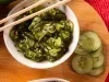 Imagem de um bowl branco sobre uma tábua de madeira com a salada de pepino em fatias e gergelim com um hashi.