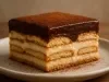 Fotografia que mostra um pedaço perfeito da torta alemã com camadas de biscoito e creme, com uma cobertura de chocolate em pó. O pedaço está sobre um prato pequeno e raso, de tom cinza claro, sobre uma mesa de madeira.