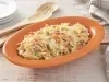 Fotografia em tons de laranja em uma bancada de madeira clara, um recipiente oval laranja com a salada de repolho, cenoura, uvas-passas e maionese. Ao lado, colheres de pau e um paninho bege xadrez.