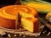 Fotografia de um bolo de milho dourado, com uma fatia cortada, revelando seu interior macio. Está sobre uma tábua de madeira, cercado por grãos de milho e espigas frescas.