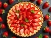 Fotografia vista de cima de uma torta de morango perfeita com pedaços decorando perfeitamente a torta com folhas de hortelã. Ao redor tem vários morangos inteiros sobre uma mesa preta.