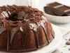 Fotografia de um bolo de chocolate, com uma cobertura brilhante que desce pelas laterais. O topo é enfeitado com raspas e granulado de chocolate, em um prato branco, com um fundo que mostra uma tigela com pedaços de chocolate.