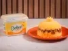 Foto de um cuscuz recheado com charque, servido em um prato laranja, ao lado de uma embalagem de margarina deline