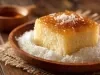 Fotografia de um bolo de milho com cobertura caramelizada, decorado com coco ralado, servido em um prato de madeira sobre um tecido rústico.