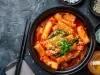 Fotografia vista de cima de um recipiente fundo preto com uma receita de Tteokbokki e cebolinha por cima. Ao lado tem dois hashis pretos sobre uma mesa preta com texturas. Do outro lado tem recipiente pequenos pretos com acompanhamentos.