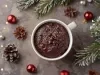 Caneca branca cheia de bolo de chocolate, decorada com gotas de chocolate e açúcar de confeiteiro. A caneca está cercada por enfeites natalinos, como ramos de pinheiro, pinhas e ornamentos vermelhos e prateados.