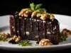 Bolo de chocolate escuro e úmido com nozes na massa, coberto por calda brilhante de chocolate meio amargo. Decorado com nozes inteiras e folhinhas de hortelã, servido de forma elegante em um prato branco.
