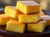 Fotografia de pedços quadrados de bolo de milho dispostos em um prato escuro. A textura é fofinha e úmida, e a cor amarela vibrante são destacadas pela iluminação.