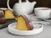 Fotografia de um pedaço de bolo em um prato branco, polvilhado com açúcar de confeiteiro, acompanhado por um garfo. Ao fundo, um bule e uma xícara de chá.
