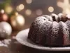 Fotografia de um bolo de chocolate em forma de bundt, em um prato branco, polvilhado com açúcar de confeiteiro. O fundo apresenta decorações natalinas em tons de ouro e prata.