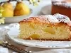 Fotografia de uma fatia de bolo de maçã em um prato branco, com uma textura leve e pedaços de fruta visíveis. O bolo é polvilhado com açúcar de confeiteiro.