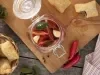 Imagem vista de cima de um pote de vidro de conserva de pimenta sobre uma tábua de madeira, com pastéis ao lado e algumas pimentas vermelhas