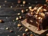 Fotografia de um bolo de chocolate coberto com uma cobertura brilhante e nozes, colocado sobre uma mesa de madeira rústica. Um fio de cobertura de chocolate escorre pelo lado.