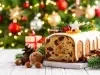 Fotografia de um bolo de frutas natalino, coberto com glacê branco e enfeitado com um biscoito em forma de estrela, frutas secas e ramos de pinheiro. Ao fundo, uma árvore de Natal iluminada e presentes.