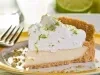 Fotografia de uma fatia generosa de torta de limão com raspas e chantilly sobre um prato de vidro com o fundo branco e a borda amarela.