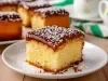 Fotografia de um pedaço de bolo quadrado com crosta dourada e interior amarelo, coberto com uma camada de chocolate brilhante e polvilhado com açúcar de confeiteiro.