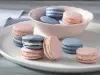 Fotografia em tons de azul e rosa em uma bancada de madeira branca clara com vários macarons coloridos em um prato branco e um paninho azul listrado.