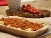 Fotografia em tons de marrom com uma travessa funda ao centro. Dentro da travessa existe algumas panquecas recheadas de carne cobertas com molho de tomate. Ao fundo existe um ramo de tomate cereja.