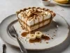 Em cima de uma mesa, prato com fatia de banoffee com massa de biscoitos crocante, doce de leite, chantilly, decorado com bananas e nozes picadas.