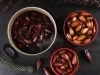 Fotografia em tom escuro vista de cima de uma mesa preta. Sobre a mesa tem uma panela cheia com água e pinhões cozidos, ao lado tem dois recipientes fundos vermelhos com mais pinhão, metade cru e metade cozido.
