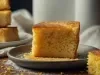 Fotografia de um pedaço de bolo de milho em um prato cerâmico cinza, com crosta dourada e textura úmida. O fundo apresenta um empilhamento de bolos semelhantes.