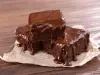 Três pedaços de brownie estão empilhados sobre papel manteiga. Eles são bem úmidos e cobertos com uma generosa camada de ganache de chocolate derretido, que escorre pelas laterais, deixando o visual ainda mais apetitoso.
