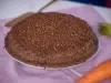 Fotografia de um bolo de cenoura com cobertura de chocolate e ascenda sobre um prato raso grande, apoiado em um pano roxo quadrado.