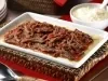 Fotografia em tons de vermelho, branco e marrom, com refratário sobre bandeja de vime com carne desfiada e molho de tomate, sobre guardanapo vermelho, ladeada por pote branco com arroz e uma colher, colher de servir, pratos, tudo sobre bancada de madeira.