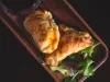 Fotografia em tons escuros vista de cima de duas empanadas recheadas com sardinha sobrepostas e apoiadas em uma tábua de madeira retangular escura.