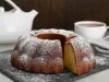 Fotografia de um bolo bundt recém-assado em uma tábua escura, polvilhado com açúcar de confeiteiro. Um bule branco e uma xícara de chá estão ao fundo.