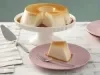 Fotografia em tons de lilás em uma bancada de madeira clara, um paninho rosa com listras cinzas, uma bailarina com o pudim com uma fatia cortada. Ao lado, um prato lilás redondo raso com uma fatia do pudim em cima dele e um garfo ao lado.