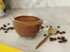 Foto aproximada de uma receita de café cremoso servido em uma xícara de vidro, com uma colher dourada ao lado e alguns grãos de café espalhados na mesa