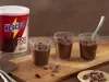 Fotografia de uma tábua retangular de madeira, pequena, com três potinhos de acrílico com o brigadeiro cremoso de colher de nescau. Ao lado tem a embalagem de NESCAU Extra Cacau 30%.