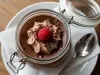 Fotografia de um pote de vidro decorativo com tampa, aberto, com a sobremesa dentro, finalizado com ganache, granulado, chantilly de chocolate e uma framboesa por cima. O pote está sobre um prato de vidro branco, raso, com um paninho branco dobrado.