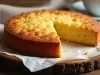 Fotografia de um bolo de milho redondo, com crosta dourada e interior macio, com uma fatia cortada, sobre papel-manteiga, em um cenário aconchegante com uma xícara desfocada ao fundo.