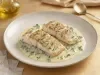 Filé de peixe branco dourado servido no molho cremoso de ervas, apresentado em prato fundo branco com acabamento sofisticado.