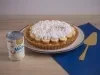 Receita de torta banoffee, servida num prato branco e decorada com chantilly e cacau em pó. A torta está ao lado de uma espátula dourada em uma bancada de madeira com um tecido na cor azul escura